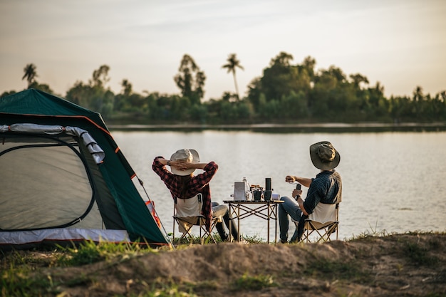 여름 방학 동안 캠핑을 하는 동안 커피 세트와 신선한 커피 그라인더를 만드는 호수 근처 텐트 앞에 앉아 휴식을 취하는 젊은 배낭 커플의 후면보기