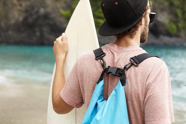 Rear view of young bearded beginner surfer wearing snapback, sunglasses and blue bag on his shoulder, holding white surfboard, looking at azure ocean water