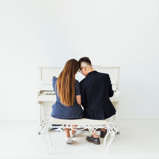 白い壁にピアノの前に座っている若いカップルの背面図
