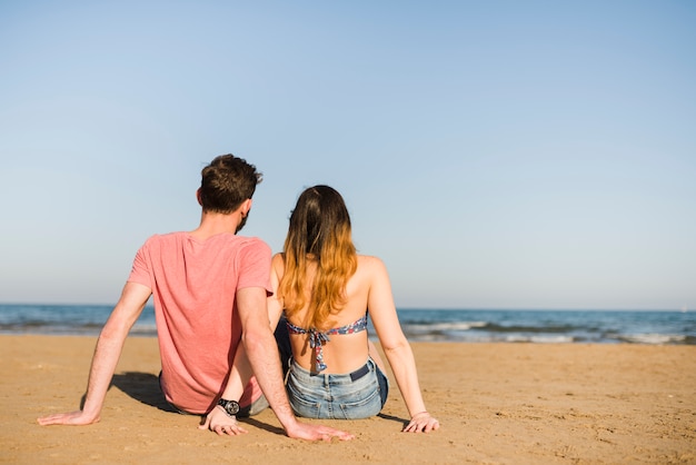 海を見て砂の上に座っている若いカップルの背面図