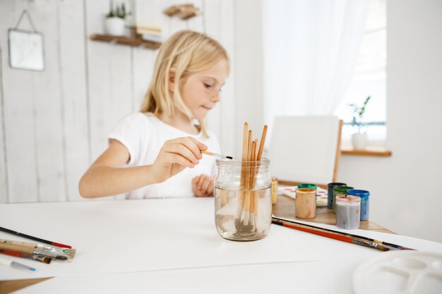 Сreative little blonde girl with freckles washing brush in jar of water during art lesson