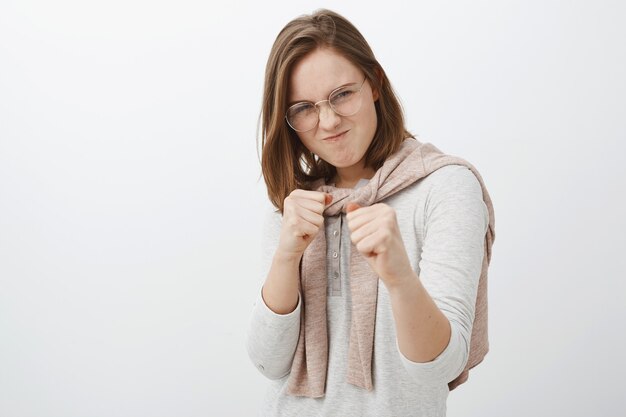 rebellious daring and playful female friend in glasses and sweatshirt tied over neck raising clenching fists and grimacing making dangerous funny expression as if fighting