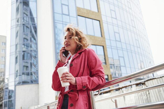 전화를 받고 있습니다. 따뜻한 빨간 코트를 입은 성인 예쁜 여자가 주말 시간에 도시를 산책했습니다.