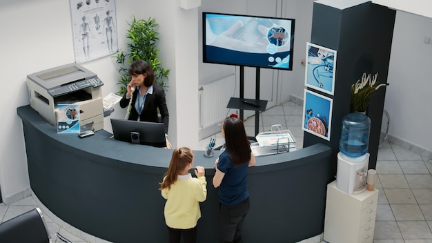 Foto gratuita receptionist che lavora nell'area di attesa dell'ospedale e offre supporto ai pazienti durante la visita di controllo, madre con bambino che ha appuntamento medico con il medico. reception presso la clinica sanitaria.