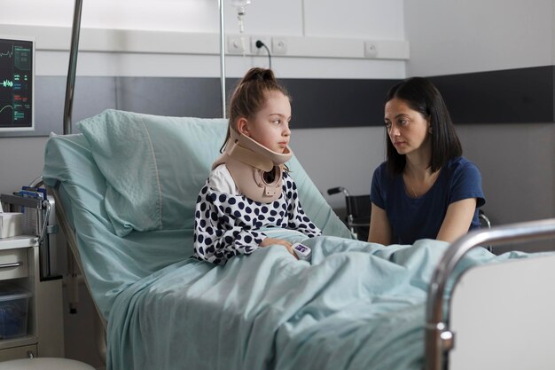 Recovering hospitalized little girl resting in recovery pediatric hospital ward while mother comforting her. Injured daughter suffering from neck pain because of car accident wearing cervical collar.