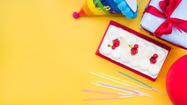 Rectangular cake with candles; balloon; gift box and paper hat on yellow background