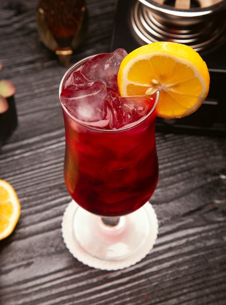 Красный алкогольный коктейль в бокале с кубиками льда и ломтиком лимона
