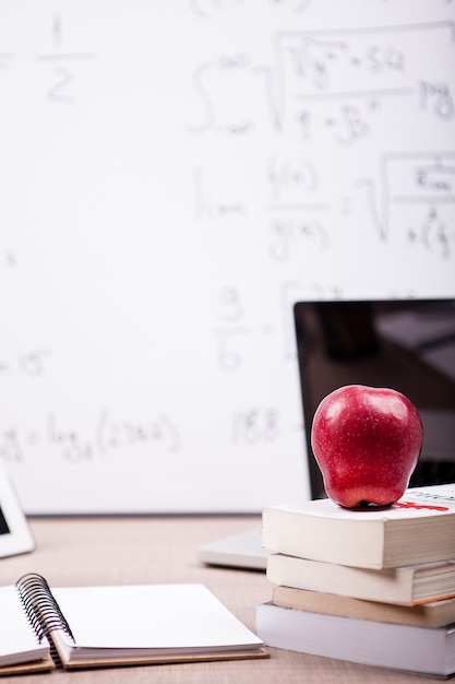 책, 공책, 연필 더미에 빨간 사과가 뒤쪽에 흐릿한 화이트 보드가 있는 탁자 위에 있습니다. 학교 개념