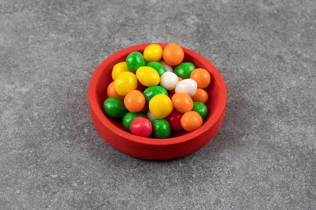 Red bowl of colorful round candies on stone table.