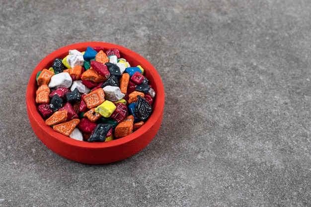 Red bowl of colorful stone candies on stone table.