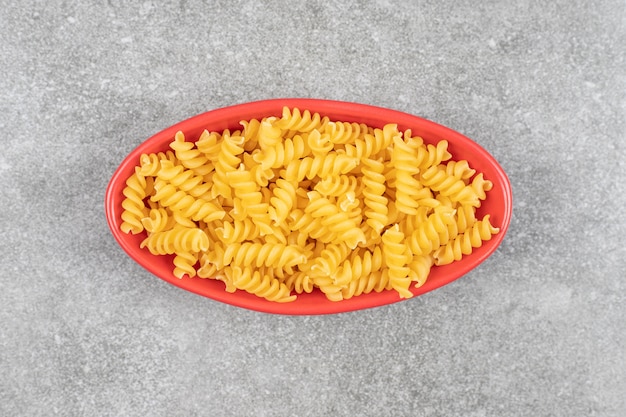 Red bowl of raw fusilli on marble surface