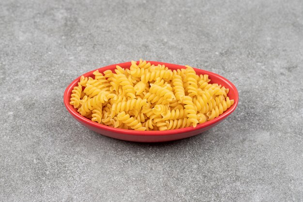 Red bowl of raw fusilli on marble surface