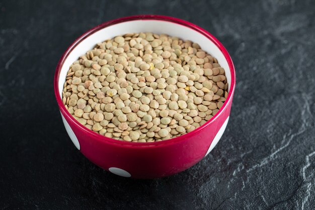 A red bowl with dried brown raw lentils on black .