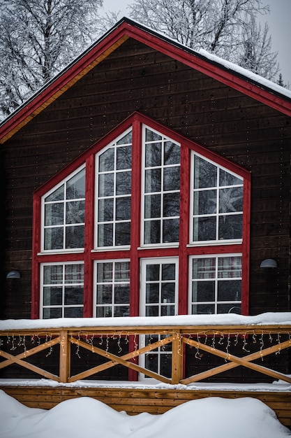 나무로 둘러싸인 숲에서 눈에 덮여 큰 창문이있는 붉은 색과 갈색 목조 주택
