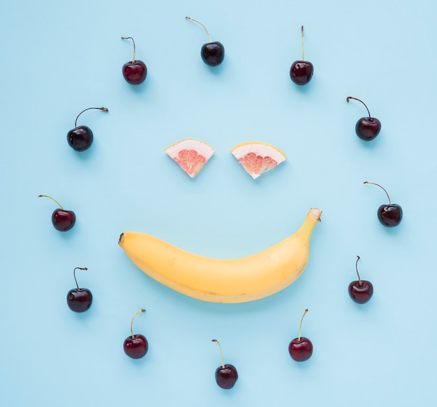 Red cherries arranged in circular frame with smiley face made with banana and grapefruit on blue background