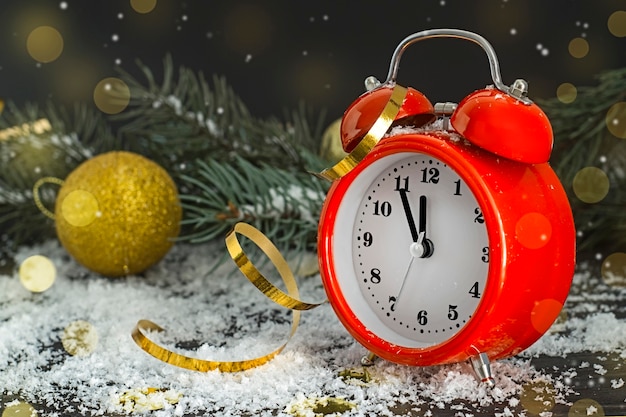 金色​の​ボケ​と​暗い​雪​の​背景​に​クリスマスツリー​の​枝​の​近く​の​赤い​時計​。​クリスマスカード​。
