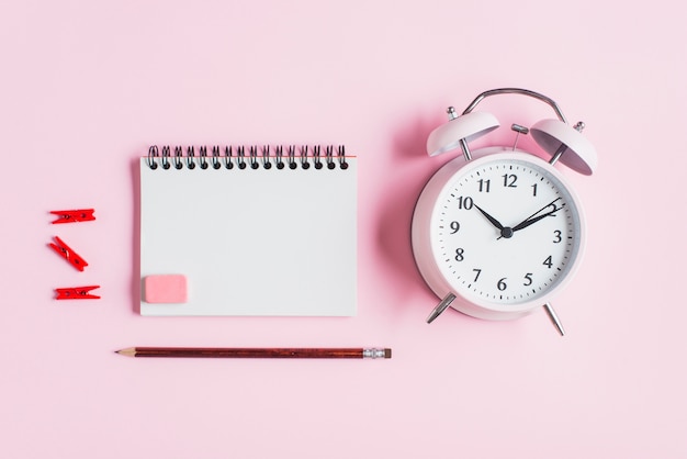 Red clothes peg; spiral notepad; rubber; pencil and alarm clock against pink backdrop