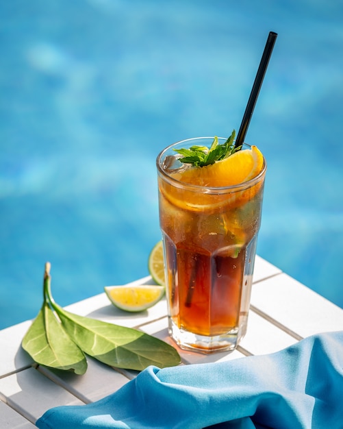Red cocktail with lemon, orange slices and hreen herbs by a pool.