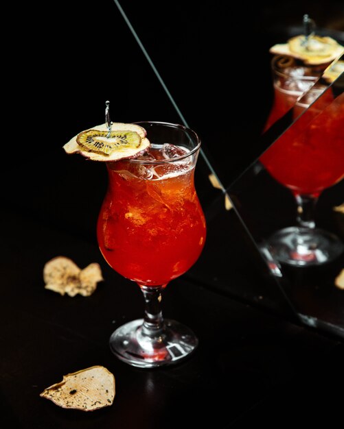 Red cocktail with minced ice cubes and kiwi slices in front of a mirror.