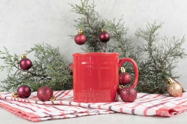 Красная кофейная чашка на скатерти с рождественскими шарами и ветвью сосны.
