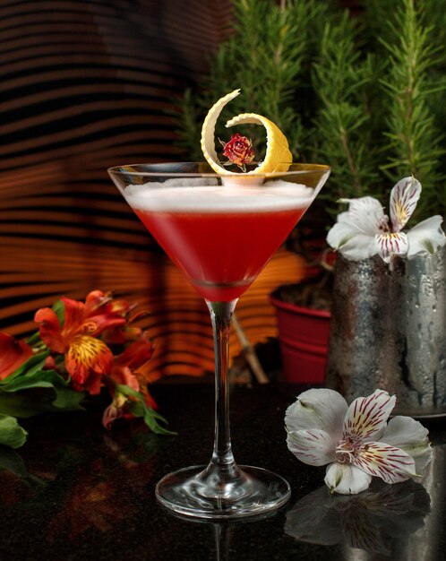 red drink in martini glass with lemon zest garnish in dim-lit bar with flowers