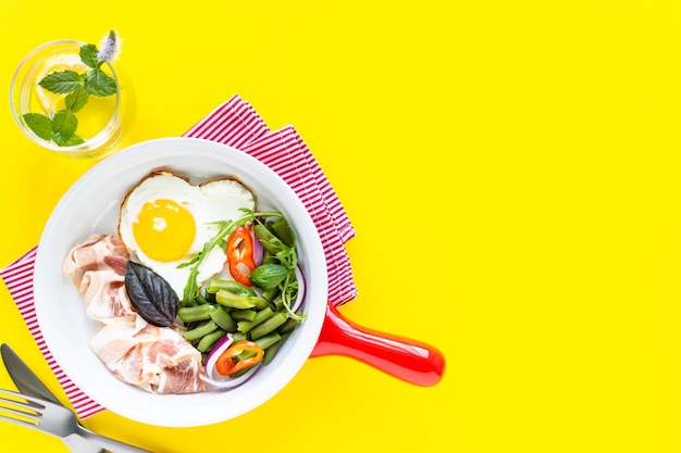 Red frying pan with delicious breakfast on a yellow, copy space. Top view, Selective focus.