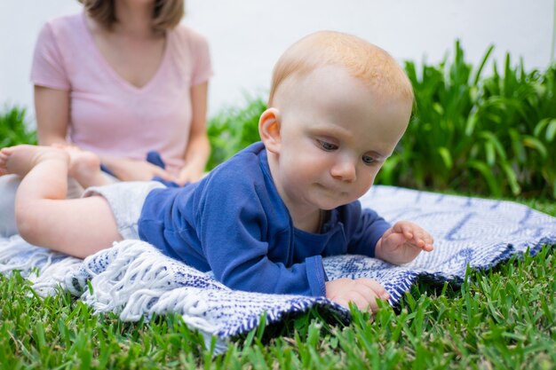 赤毛の女の赤ちゃんは格子縞の上に横たわって、興味を持って草に触れ、よそ見します。庭のポートレート、クローズアップ。座っている若いお母さん。夏の家族の時間、晴れた日、新鮮な空気の概念