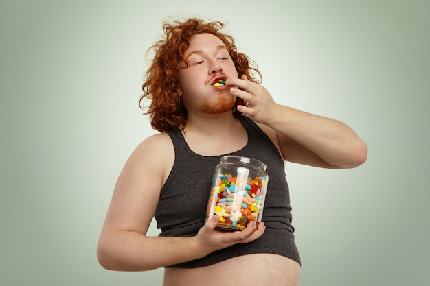 red haired Caucasian overweight man wearing undersized tank top looking ridiculous while eating marmalades out of glass jar, keeping stuffing them into his mouth. Obesity and gluttony