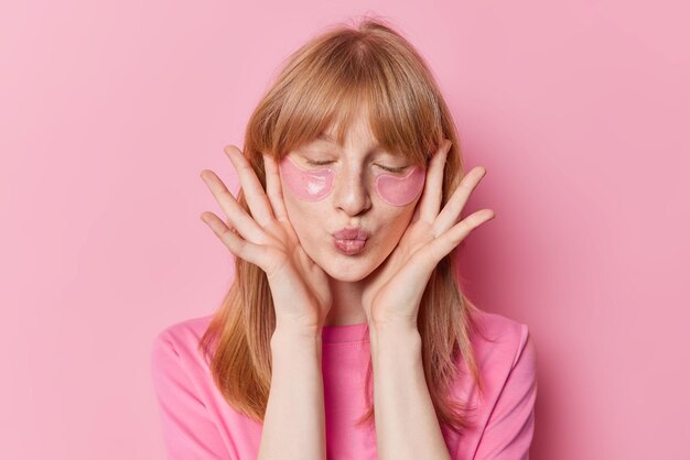 Red haired freckled girl keeps lips folded has freckled skin applies hydrogel patches under eyes undergoes beauty procedures wears casual t shirt isolated over pink background Facial treatment