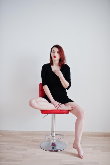 Red haired girl on black dress tunic sitting on red chair against white wall at empty room