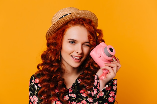 花柄のブラウスとブラウスの赤い髪の少女がウィンクし、オレンジ色のスペースにピンクのカメラを持っています。