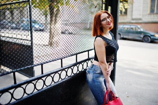 Red haired girl in glasses with red handbag posed at street of city