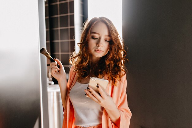 Red-haired girl making makeup in bathroom. Photo of serious ginger woman posing with phone at home.