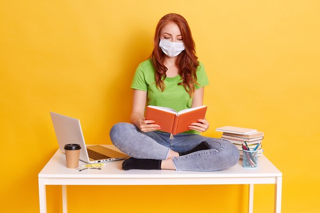 Red haired girl in medical mask sits on table with computer and books, reading, looks concentrated