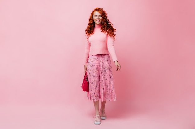 Free Stock Photo: Red-haired girl in pink skirt and sweater posing with bright bag on isolated wall