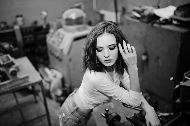 Red haired girl wear on short denim shorts and white blouse posed at industrial machine at the factory