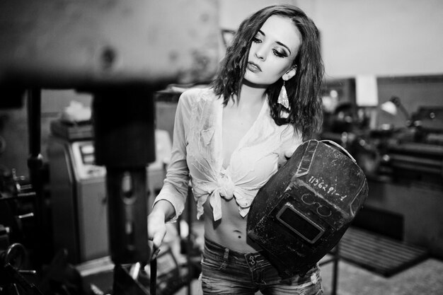 Red haired girl wear on short denim shorts and white blouse with welding mask at hands posed at industrial machine at the factory