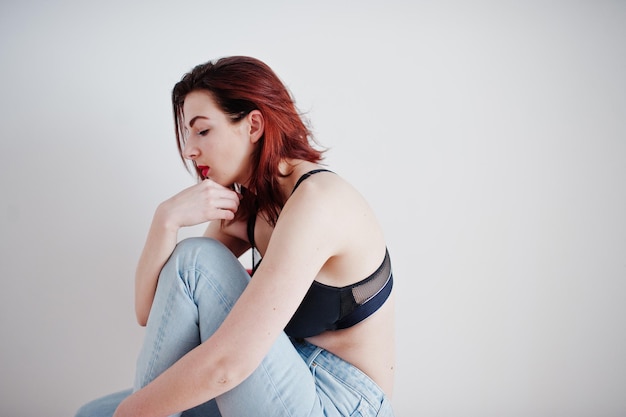 Red haired girl with a big bust on black bra and jeans against white wall at empty room