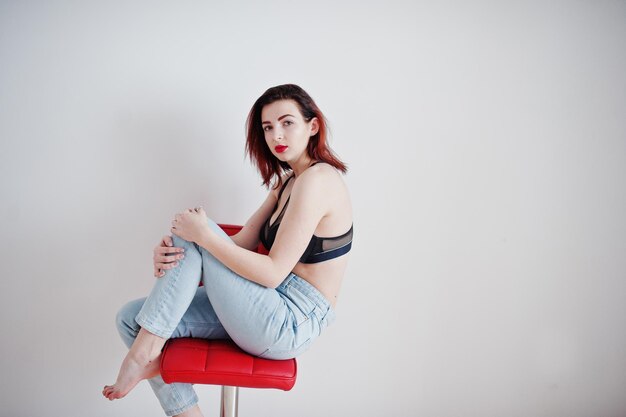 Red haired girl with a big bust on black bra and jeans sitting on red chair against white wall at empty room