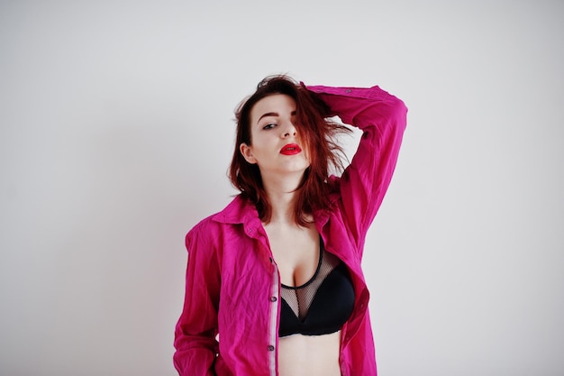 Red haired girl with a big bust on black bra and rose shirt against white wall at empty room