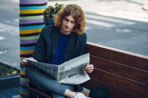 Red haired hipster man sitting on bench reading newspaper