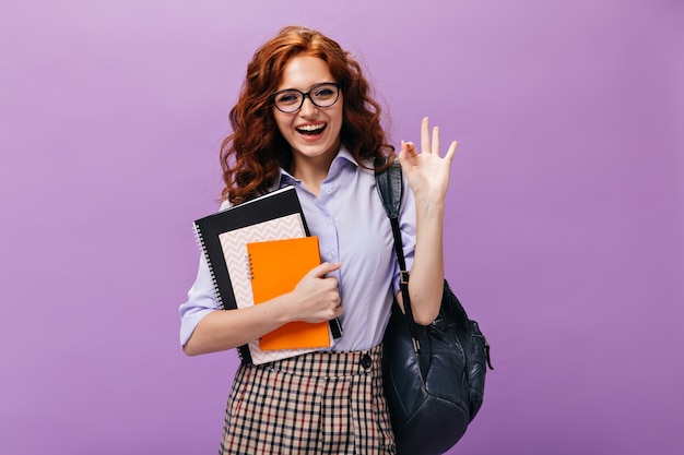 Red haired lady in eyeglasses holds books and shows ok sign