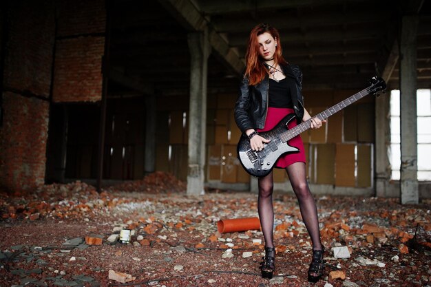 Red haired punk girl wear on black and red skirt with bass guitar at abadoned place Portrait of gothic woman musician