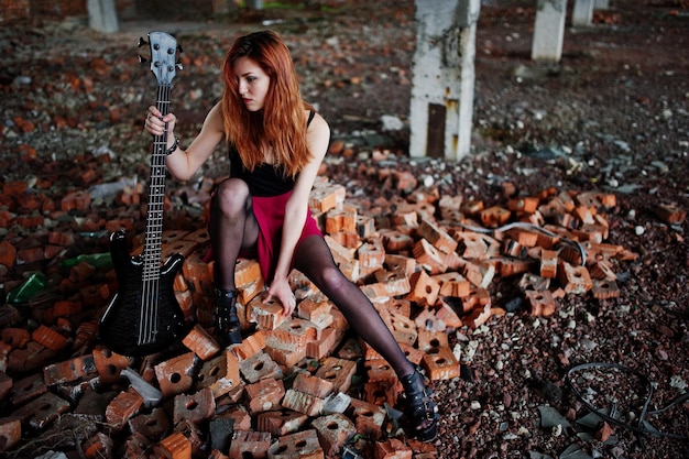 Red haired punk girl wear on black and red skirt with bass guitar at abadoned place Portrait of gothic woman musician