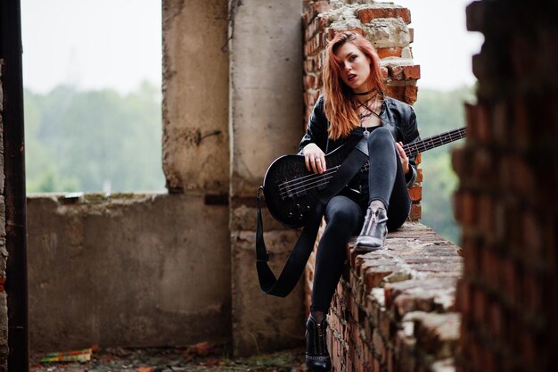 Red haired punk girl wear on black with bass guitar at abadoned place Portrait of gothic woman musician
