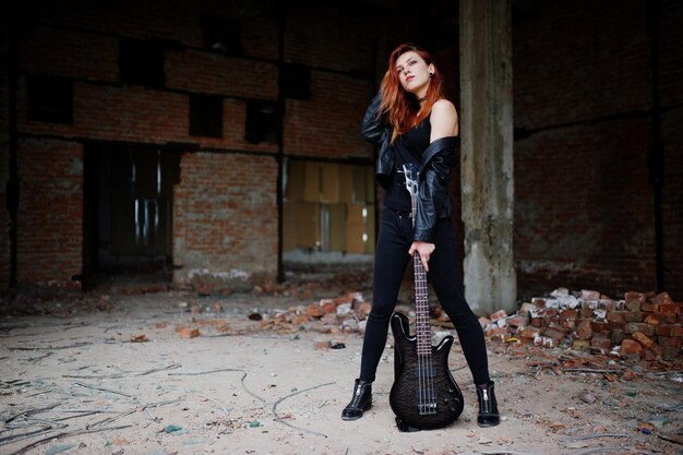 Red haired punk girl wear on black with bass guitar at abadoned place Portrait of gothic woman musician