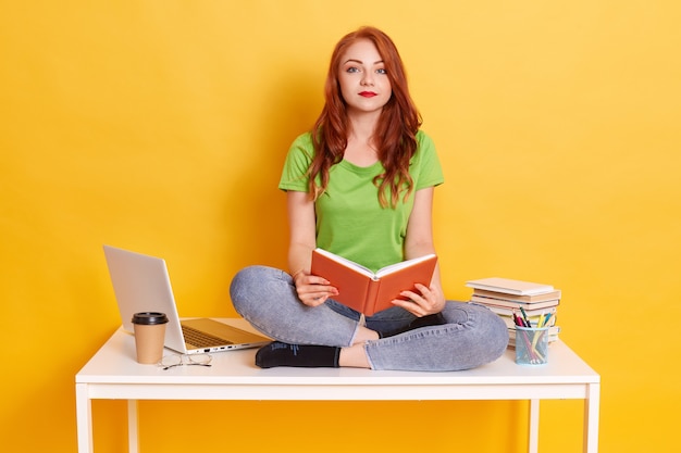 Red haired student girl sitting on table with crossed legs, holding book in hands, looks at camera, being tired from distance learning, wearing casual clothing.