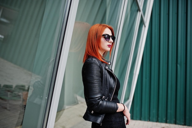 Free photo red haired stylish girl in sunglasses wear in black against large windows street fashion portrait