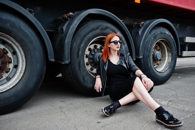 Рыжая стильная девушка в черном сидит на фоне больших колес грузовика