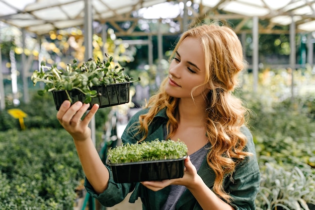 Red-haired sweet lady intently examines evergreen small plants. Closeup portrait of model of European appearance in garden.